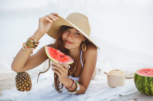 Beautiful woman holding watermelon and resting on the beach near the water. Tropical fruits diet concept. Summer holiday idyllic. Portrait of happy young slim mixed race Asian Caucasian girl in bikini and hat wearing jewelry chilling at sea eating sweet food.