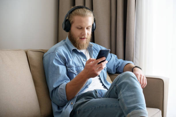 il giovane che indossa cuffie wireless con in mano la cella ascoltando musica, audiolibri, guardando video mobili, imparando la lingua o effettuando videochiamata sullo smartphone in applicazione si siede sul divano di casa. - head in hands audio foto e immagini stock