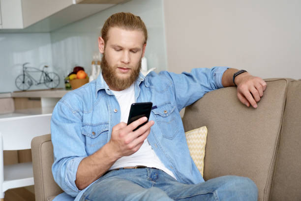 istock Serious young bearded man user holding smartphone texting message using online education app sit on sofa at home. Millennial guy using cellphone applications surfing web, read news on phone indoors. 1221338489