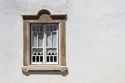 Bright white wall of a house during a bright sunny day. Window with white wooden details and old fashioned curtains. Stone frame. Estoi, Portugal.