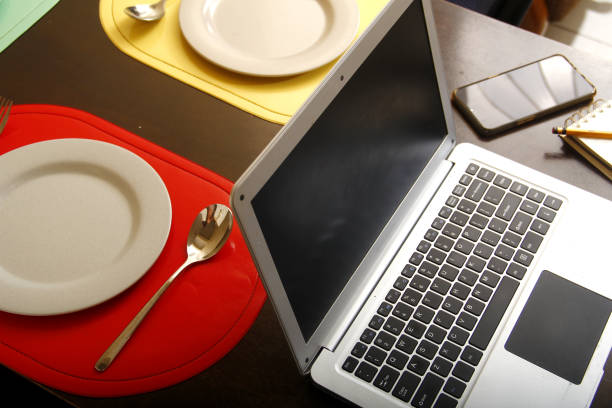istock Laptop computer, smartphone, notebook, pencil, plates and utensils on a dining table 1221340899