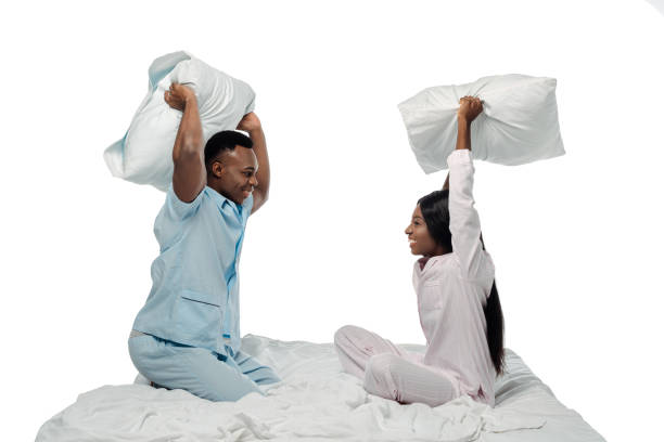 pareja afroamericana feliz tener pelea de almohada en la cama en pijama aislado en blanco - bed couple pillow fight men fotografías e imágenes de stock