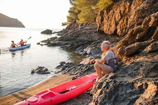 Side view of senior male in mid 70s wearing life jacket and sitting on shoreline rocks enjoying morning sunshine as friends paddle by in tandem kayak.