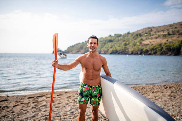 Front view of Spanish paddleboarder in early 40s standing on beach and smiling at camera before exercising in Mediterranean.