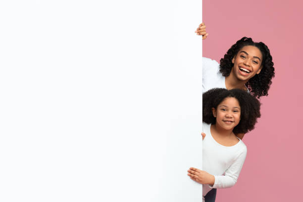Cheerful Black Mom And Daughter Peeking Out Behind White Advertisement Board