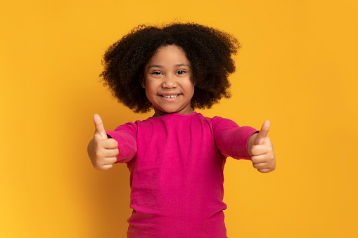 Like it. Cute little black girl showing thumbs up gesture and smiling at camera, posing over yellow studio background with free space