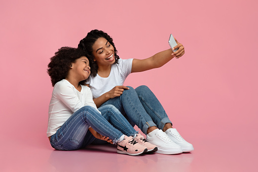 Family Selfie. Cheerful black mother taking photo with her little cute daughter, posing together on floor over pink studio background, free space