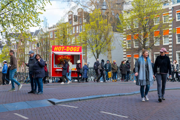 Hot dog stand on a bridge on a bright spring day in Amsterdam, Netherlands. There are tourists walking over the bridge and queueing at the stand with a young couple in the foreground.