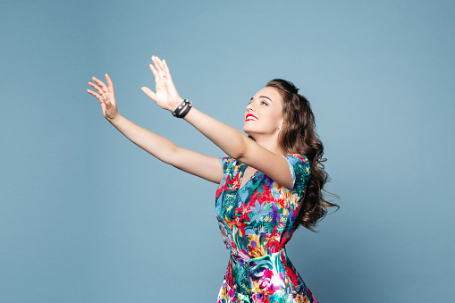 Studio portrait of beautiful happy brunette lady in summer floral dress pulling her arms up in the air to get something. Isolate on blue background.