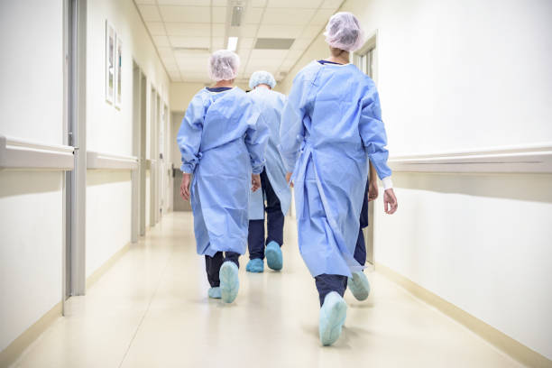 Team of medical staff in personal protective equipment walking in hospital corridor Rear view of male and female surgical team wearing gowns, caps, shoe covers, and walking down hospital corridor to operating room. sydney photos stock pictures, royalty-free photos & images