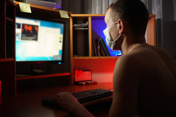 istock Man with a bare torso in a medical mask sits at a computer and studies information in a dark room. Self-isolation 1221365319