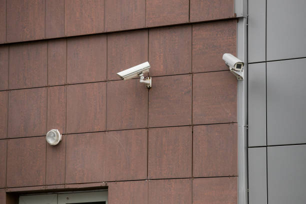 istock Big white surveillance camera on light beige stone column against background of public building with blue window; close-up 1221373235