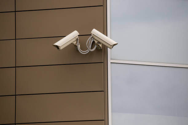 istock Big white surveillance camera on light beige stone column against background of public building with blue window; close-up 1221374310