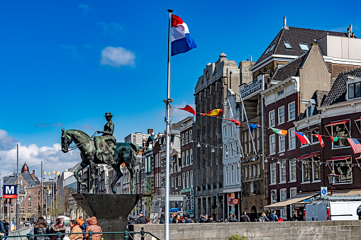 Amsterdam, Netherlands-April ,2019: Statue of Queen Wilhelmina, with crowds of people and tourists on the street and a Dutch flag in the foreground.