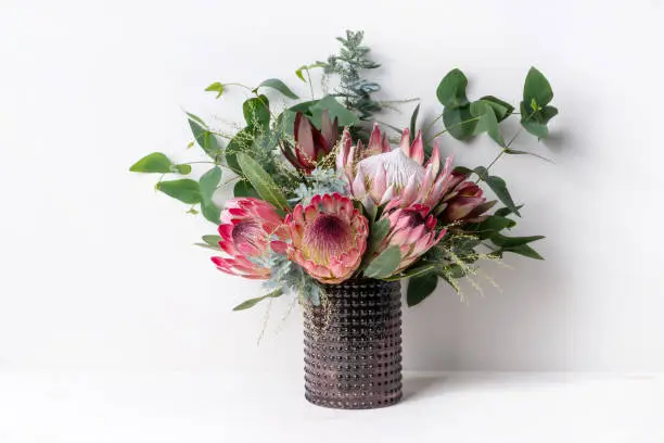 A elegant floral arrangement in a rustic brown vase on a table with a white background. Flower bunch includes a pink king protea, pink ice proteas, leucadendrons, wattle leaves and eucalyptus leaves.