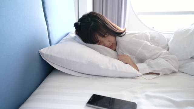 Asian woman waking up and sleep with smartphone alarm clock her snoozing at morning in hotel room travel tourist concept 4k resolution