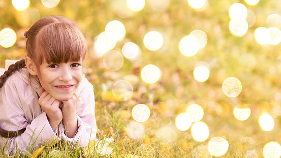 Little girl lying at park. Happy kid at lights bokeh background. Autumn action. Stop quarantine. Yellow grass. Miracle backdrop. Party outdoor. Horizontal banner with copyspace