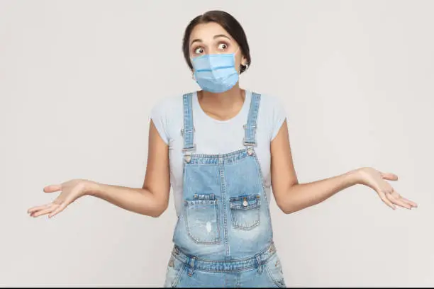 I don't know. Portrait of thoughtful young brunette woman with surgical medical mask in denim overalls standing raised arms and looking away. indoor studio shot isolated on gray background.