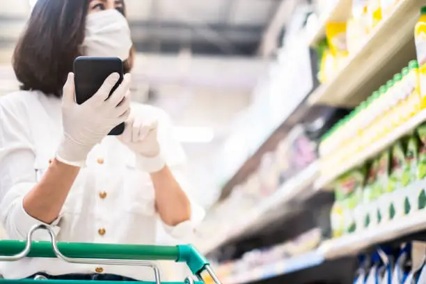 Asian woman wearing face mask and rubber glove push shopping cart in supermarket department store. Girl hold smartphone choose & look grocery things to buy during coronavirus crisis, covid19 outbreak.