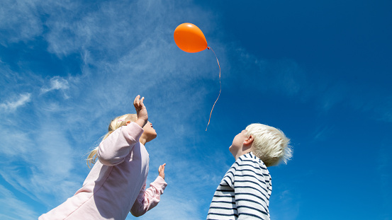 Low angle view of boy and girl releasing orange balloon in blue sky.