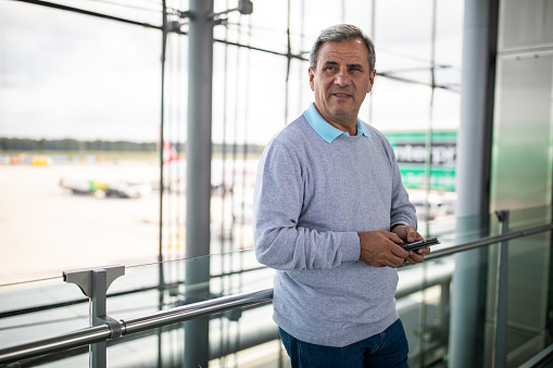 Mature casually clothed gray haired man standing, waiting for a plane and holding a mobile phone at airport station