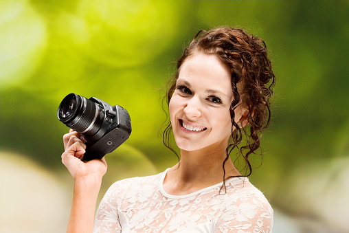 Portrait of aged 20-29 years old who is beautiful with curly hair caucasian female tourist in front of defocused background wearing dress who is showing cool attitude who is photographing and holding camera