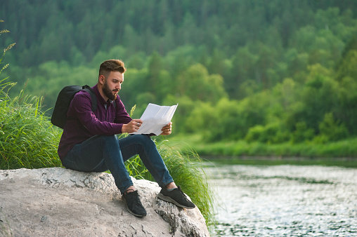 A bearded tourist man, on the shore of a mountain river, is studying a map