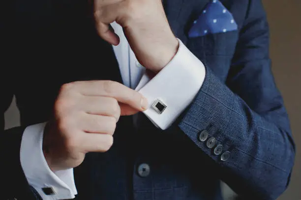 Elegant fashion man looking at his cufflinks while fixing them. Handsome groom dressed in black formal suit, white shirt and tie is getting ready for wedding.