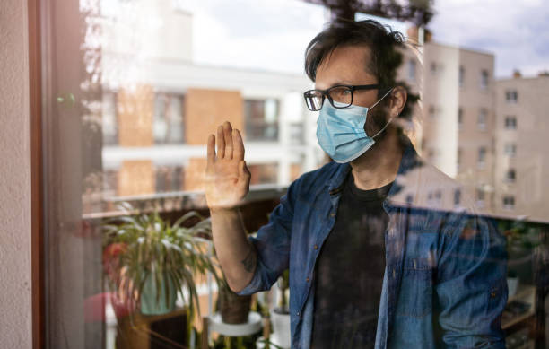 Man with face mask looking out of window Man in isolation at home for virus outbreak stay at home order stock pictures, royalty-free photos & images