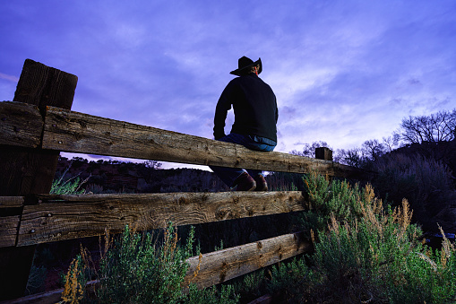 Country Western Cowboy Rancher Man at Sunset on Fence - Rancher reflecting and pondering a hard day's work with views of dusk sky and silhouetted iconic outline.