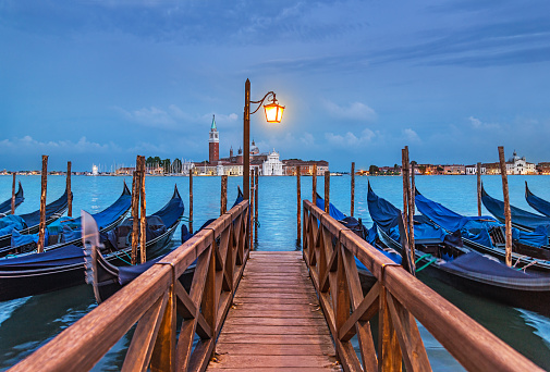 Evening Venice seascape with gondolas at moorings, light of lantern at wooden bridge, view at famous San Giorgio Maggiore island with basilica and campanile from Piazza San Marco. Italy, Europe