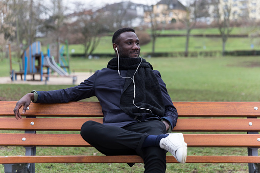 Young black man listening to audiobook seated on park bench and looking away.