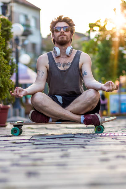 Hippie fashion Man doing yoga, relaxing on skateboard Big picture of hipster young man doing yoga, relaxing and sitting on a skateboard on the street. World Skateboarding and Yoga Day. The concept of freedom and a healthy lifestyle in the sport of yoga and skateboarding man yoga tattoo stock pictures, royalty-free photos & images
