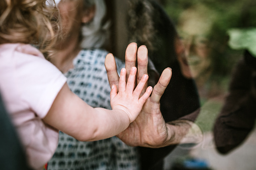 A mother stands with her daughter, visiting senior parents but observing social distancing with a glass door between them.  The granddaughter puts her hand up to the glass, the grandfather and grandmother doing the same.  A small connection in a time of separation during the Covid-19 pandemic.