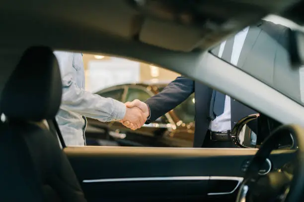 Photo of Buyer of car shaking hands with seller in auto dealership, view from interior of car.