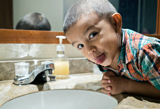 Latino boy of average age of 6 years old dressed casually is leaning on the sink where he wants to wash his hands but looks at the camera and sticks his tongue out