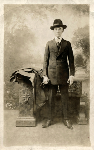 Early 1900 portrait of a young man in a suit and Fedora hat holding a cigar in one hand and gloves in the other.