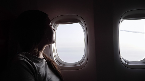 woman in grey t-shirt silhouette against airliner bright windows resting and enjoying in passenger cabin closeup