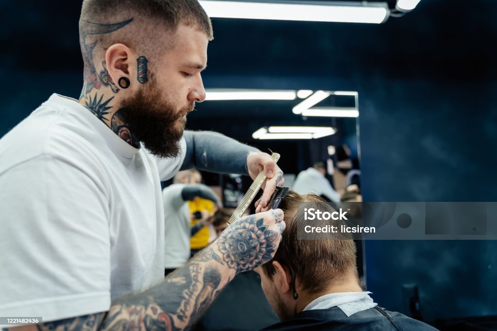 Professional tattooed barber using cutthroat razor cutting hair. Attractive male is getting a modern haircut in barber shop. Close up Professional barber with open cutthroat razor shaves hair on man's temples. Adult Stock Photo