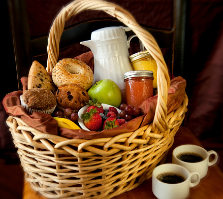 Wicker basket containing muffins, bagels, scones, strawberries, banana, carafe of coffee, two mason jars of orange juice and grapefruit juice. Basket is sitting next to two white mugs full of coffee.