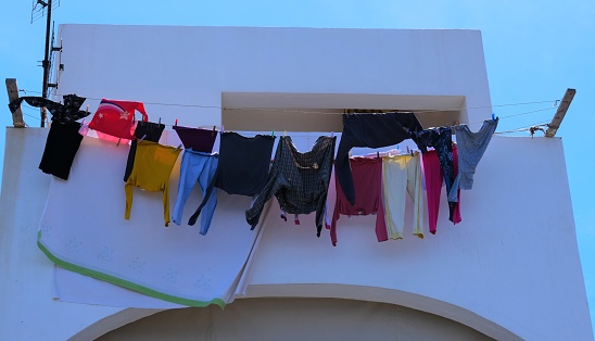 Laundry hanging out to dry in Greece.  Hanging from patio's, balconies and from the side of buildings.  Clothes lines constructed from various materials and clothes, sheets and towels hanging in the sun to dry.  Whitewashed buildings, narrow alleyways and stone buildings against a blue sky.