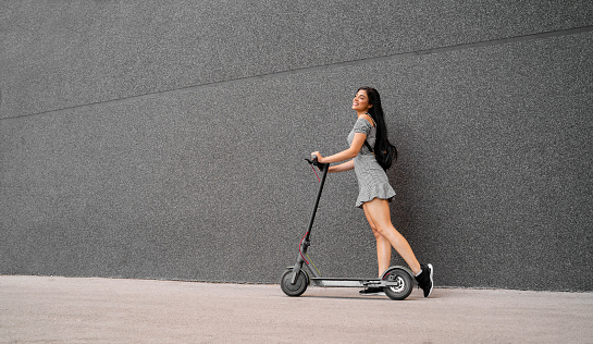 Happy young woman with e scooter smiling next to black wall, copy space