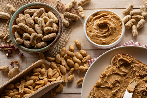 Peanut composition with organic raw unshelled peanuts, roasted peanuts, peanut butter bowl and one slice of bread with peanut butter.