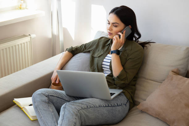 istock Beautiful young woman using laptop and talking on cellphone at home 1221504322