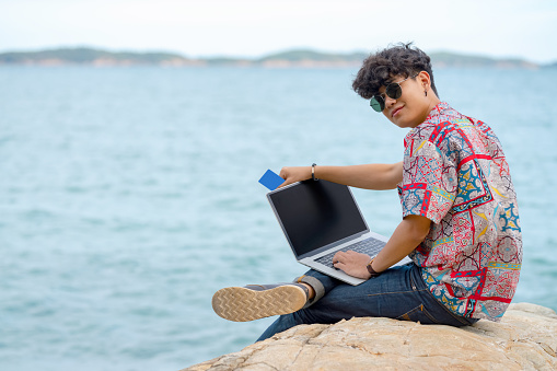 Smiling young smart Asian man guy sitting on rocky seacoast beach in summer vacation using laptop computer with internet for outdoors working and online shopping with credit card for e-banking.