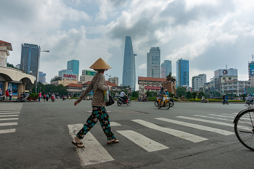 Saigon Vietnam October 12 2013; Woman in traditional Asian style conical hat crosses road in a busy city intersection surrounded by conflicting low buildings and modern high-rise.