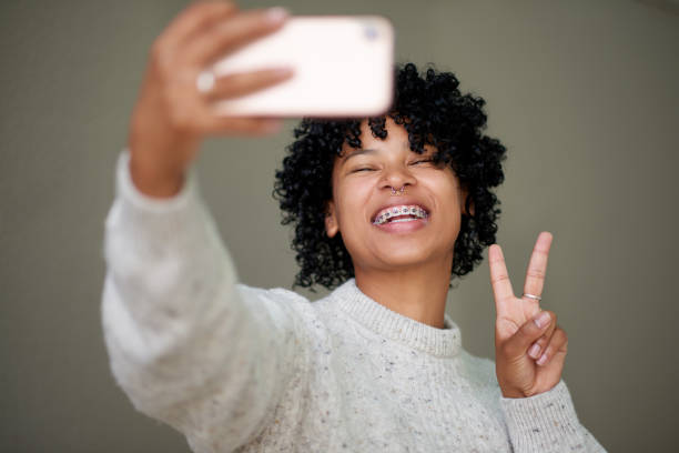 Studio shot of a young woman making a peace sign with her fingers while taking selfies against a grey background