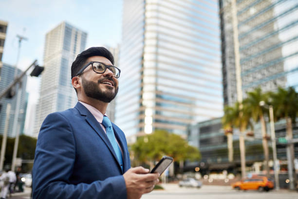 Portrait of Optimistic Hispanic Businessman with Smart Phone Low angle view of bearded and bespectacled Miami businessman holding smart phone and looking up with hopeful expression. businesswear stock pictures, royalty-free photos & images