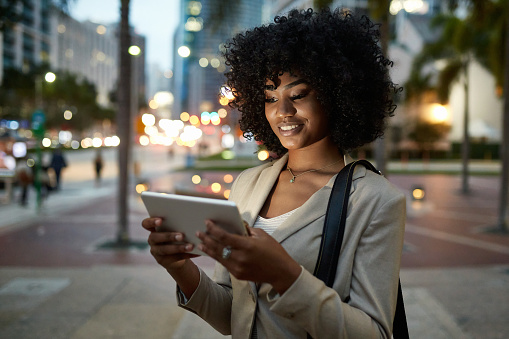 Three-quarter front view of 21 year old African-American corporate professional standing outdoors in downtown Miami and reading digital tablet in early evening.
