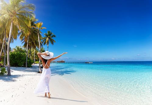 A woman with white hat walks down a tropical paradise beach with palm trees and turquoise sea
